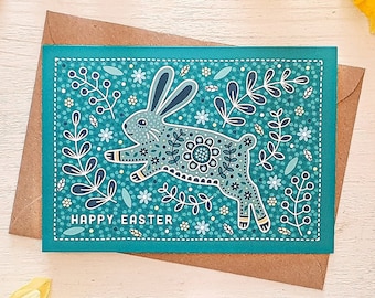 Peut inclure: Une carte de vœux rectangulaire sur fond sarcelle représentant un lapin bondissant aux contours blancs, entouré de motifs floraux et de feuilles. Le texte "HAPPY EASTER" est imprimé en blanc dans le coin inférieur gauche de la carte posée sur une enveloppe brune.