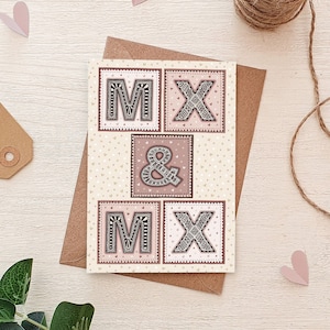 May include: A white card with a pink and white patterned background. The card has four squares with the letters "M" and "X" in each square. The letters are in a black and white patterned design. The word "&" is in the center square. The card is perfect for a wedding or engagement.