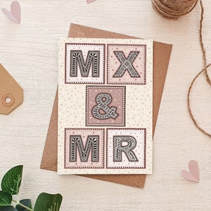 May include: A card with a cream background and pink and white polka dots. The card has four squares with the letters "M", "X", "&", and "M" and "R" in black with a decorative pattern. The letters are surrounded by a pink border.