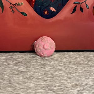 May include: A pink, round bath bomb with glitter accents. The bath bomb is on a gray and white textured surface, with a red and floral patterned background.