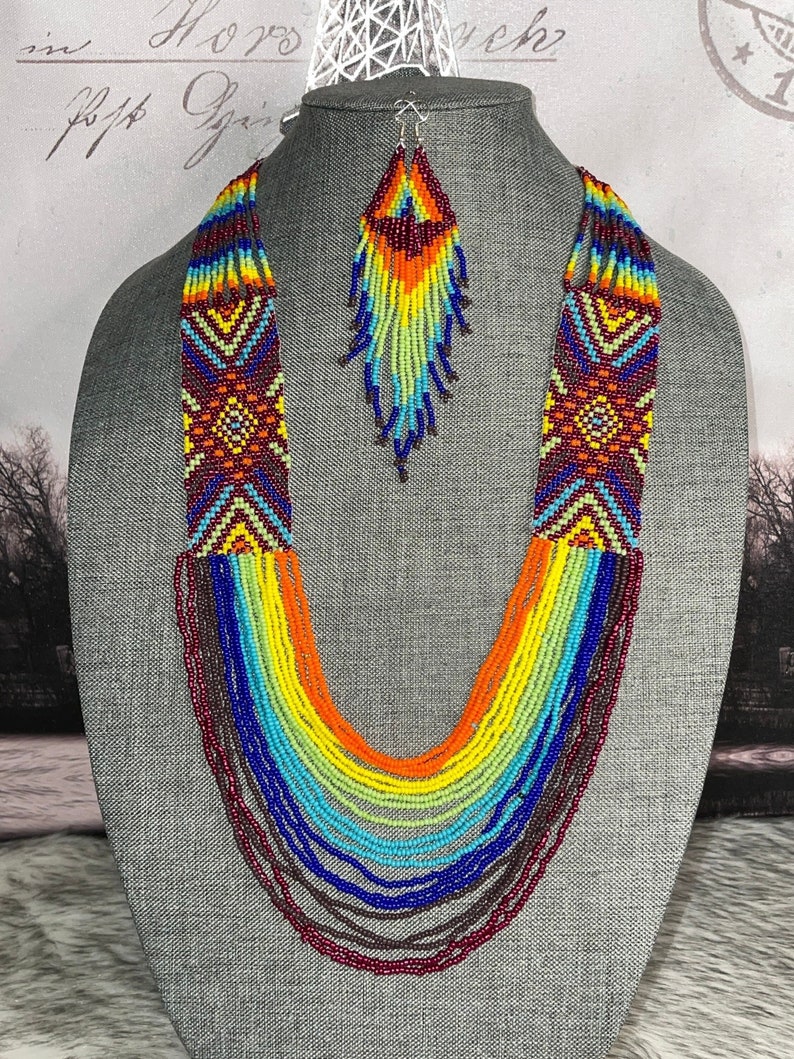 Op de afbeelding: Een meerrijige kralenketting en bijpassende oorbellen. De ketting is gemaakt van kleurrijke kralen in tinten rood, oranje, geel, groen, blauw en paars. De oorbellen zijn lang en hangend, met een vergelijkbaar kleurenschema als de ketting.