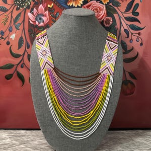 Op de afbeelding: Een meerrijige kralenketting met een geometrisch patroon in tinten roze, geel, groen en wit. De ketting wordt getoond op een grijze mannequin.