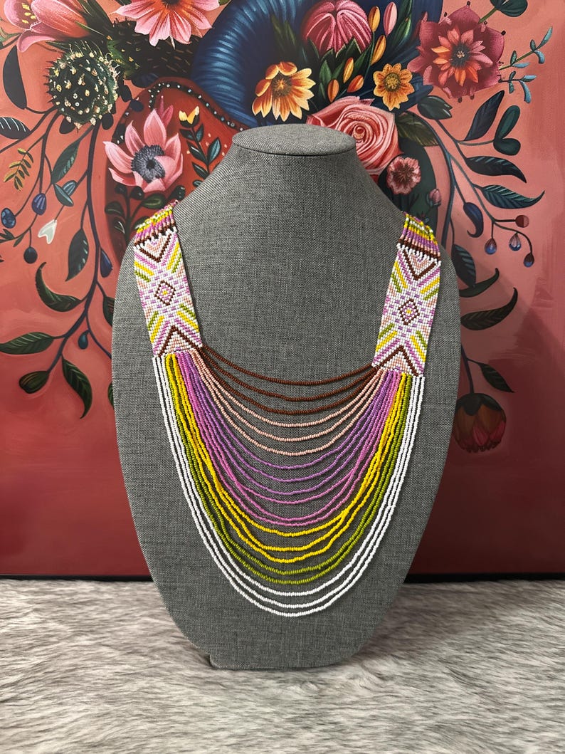 Op de afbeelding: Een meerrijige kralenketting met een geometrisch patroon in tinten roze, geel, groen en wit. De ketting wordt getoond op een grijze mannequin.