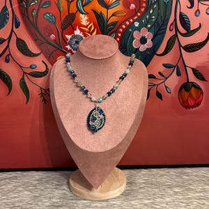 Puede incluir: Collar de cuentas con colgante de lágrima, expuesto en un expositor rosa. El collar tiene cuentas azules, verdes y transparentes, con un colgante azul y plateado. El fondo es una pintura floral. El expositor está sobre una base de madera.