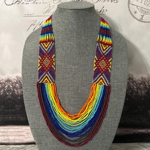 Op de afbeelding: Een meerstrengs kralenketting met een kleurrijk regenboogpatroon. De ketting is gemaakt van kleine, ronde kralen in tinten rood, oranje, geel, groen, blauw en paars. De kralen zijn in een aflopend patroon gerangschikt, met de grootste kralen bovenaan en de kleinste kralen onderaan. De ketting wordt gepresenteerd op een grijze mannequin.