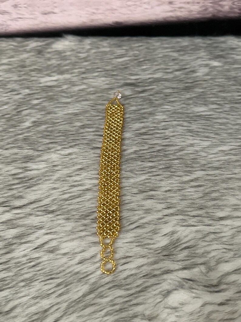 Puede incluir: Un brazalete de cadena dorada con un dise&ntilde;o delicado y tejido. El brazalete est&aacute; hecho de peque&ntilde;os eslabones redondos que est&aacute;n estrechamente tejidos.
