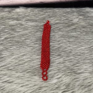 Puede incluir: Un brazalete rojo de cuentas con un dise&ntilde;o sencillo. El brazalete est&aacute; hecho de peque&ntilde;as cuentas redondas que est&aacute;n enhebradas en una l&iacute;nea recta. El brazalete mide aproximadamente 18 cm de largo y tiene un cierre en el extremo.