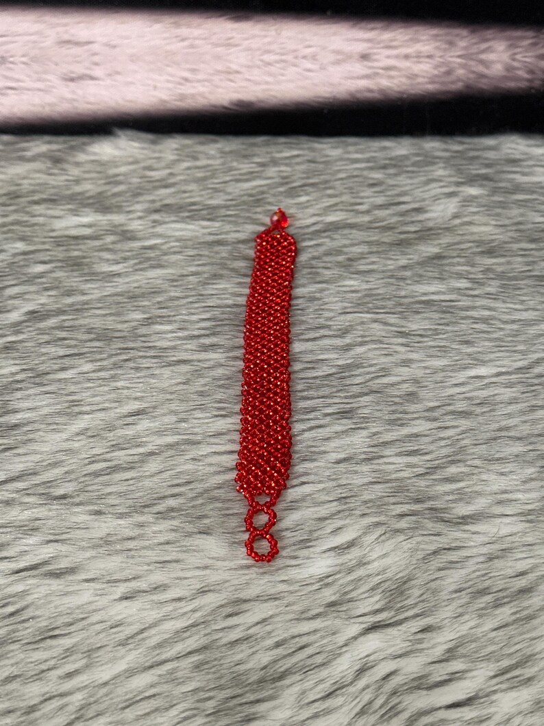 Puede incluir: Un brazalete rojo de cuentas con un dise&ntilde;o sencillo. El brazalete est&aacute; hecho de peque&ntilde;as cuentas redondas que est&aacute;n enhebradas en una l&iacute;nea recta. El brazalete mide aproximadamente 18 cm de largo y tiene un cierre en el extremo.