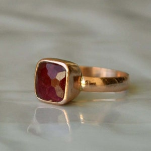 Puede incluir: Un anillo de oro rosa con una piedra preciosa cuadrada, facetada, de color rojo oscuro.