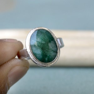 May include: A silver ring featuring a large, oval-cut emerald gemstone. The emerald has a deep green color and is set in a silver band with a textured border. The ring is held up by a hand.