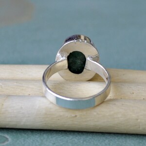 May include: A silver ring with an oval, dark green gemstone. The ring's band is smooth and polished, and the gemstone is set in a bezel setting. The ring is displayed on a light-colored wooden surface.
