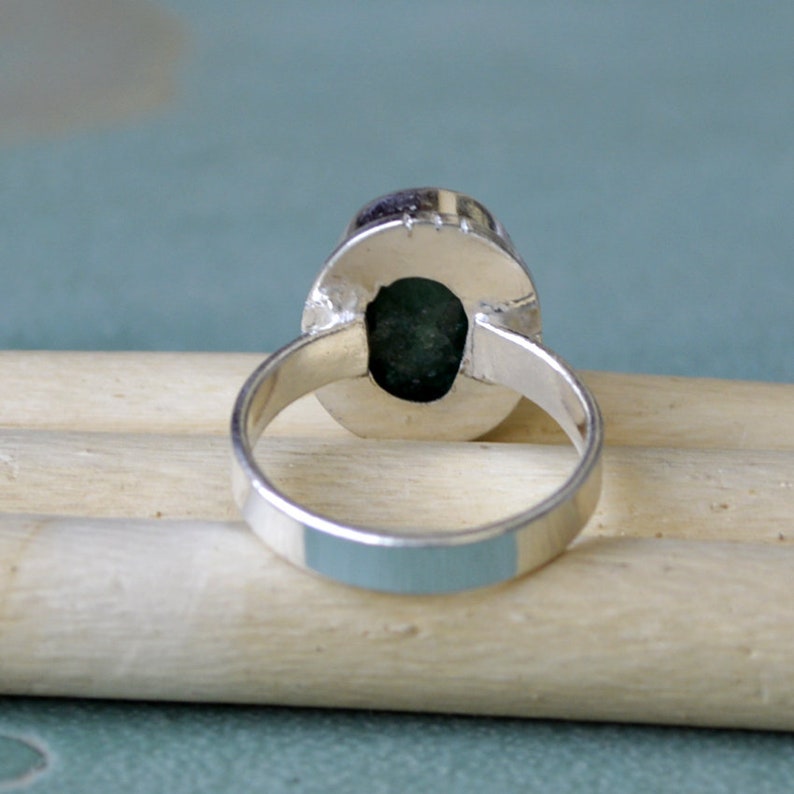 May include: A silver ring with an oval, dark green gemstone. The ring's band is smooth and polished, and the gemstone is set in a bezel setting. The ring is displayed on a light-colored wooden surface.