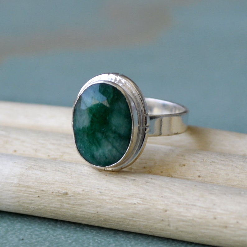 May include: A silver ring featuring a large, oval-cut emerald gemstone. The emerald has a deep green color and is set in a bezel setting. The ring band is a simple, polished silver. The ring is displayed on a light-colored wooden surface.