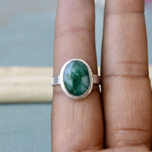 May include: A silver ring featuring an oval-cut, deep green gemstone. The gemstone is set in a bezel setting, and the band is a simple, polished silver. The ring is shown on a finger, highlighting the jewelry's design and color.