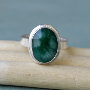 May include: A silver ring featuring a large, oval-cut, deep green gemstone. The gemstone is set in a bezel setting with a textured band. The ring is displayed on a light-colored wooden surface against a soft blue background.