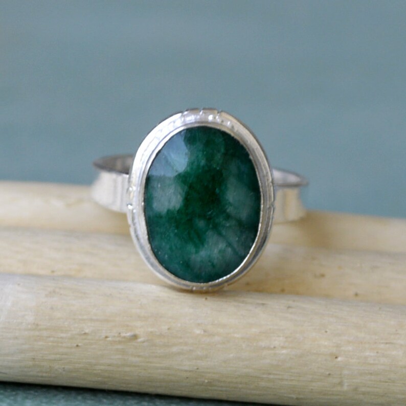 May include: A silver ring featuring a large, oval-cut, deep green gemstone. The gemstone is set in a bezel setting with a textured band. The ring is displayed on a light-colored wooden surface against a soft blue background.