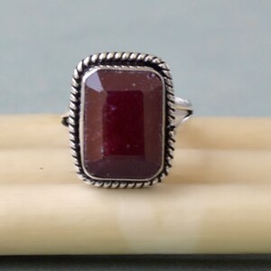 May include: A silver ring with a dark red rectangular gemstone set in a rope-like border.
