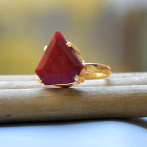 May include: A gold ring featuring a large, faceted, red gemstone. The gemstone is set in a prong setting and has a pointed shape.