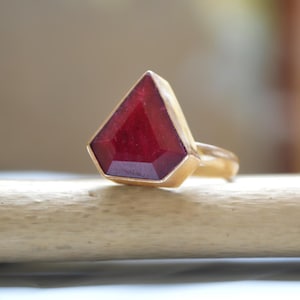 Könnte beinhalten: Ein goldener Ring mit einem großen, facettierten, rubinroten Edelstein. Der Edelstein ist in einer goldenen Fassung gefasst und hat eine spitze Form.
