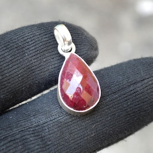 May include: A teardrop-shaped pendant with a faceted, deep red gemstone set in a silver-colored metal. The pendant has a loop at the top for a chain. The gemstone reflects light, showcasing its cut and color.