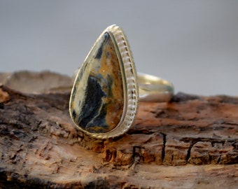 May include: A silver ring featuring a teardrop-shaped stone. The stone is a blend of brown, black, and white colours. The ring is displayed on a piece of wood.