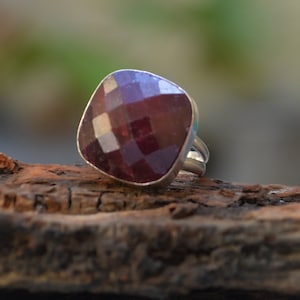 May include: A silver ring with a large, faceted, square-shaped, deep red gemstone. The gemstone has a multi-faceted surface that reflects light. The ring is set on a piece of wood.