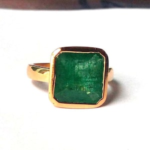 Puede incluir: Un anillo de oro con una gran esmeralda de corte cuadrado. La esmeralda es de color verde intenso y está engastada en un bisel de oro. La banda del anillo también es de oro, ofreciendo un diseño clásico y elegante.