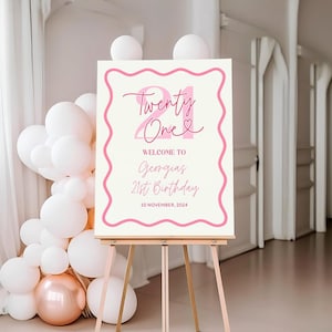 May include: A pink and white welcome sign with the text "Twenty One" in pink script with a heart symbol. The sign also includes the text "Welcome to Georgia's 21st Birthday 10 November 2024". The sign is on a wooden easel.