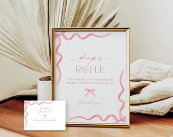 May include: A pink and white sign with a gold frame that says "diaper raffle" with pink ribbons around the border. The sign is on a wooden table with a white blanket and a dried palm leaf plant.