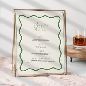 May include: A bar menu sign with a gold frame and a cream-colored background. The menu lists wine, beer, spirits, and signature cocktails. The text is in a handwritten style with a green wavy border. Two cocktails sit on a wooden tray.