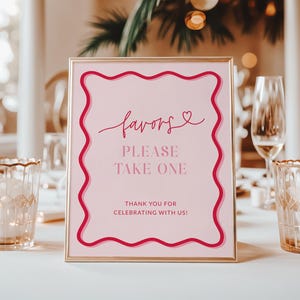 Könnte beinhalten: Ein gerahmtes rosa Schild mit dem Wort "favors" in Schreibschrift und dem Text "PLEASE TAKE ONE" in einer Serifenschrift. Das Schild hat einen wellenförmigen, dunkelrosa Rand und steht auf einem weißen Tisch. Goldgerahmte Kerzenhalter und Weingläser sind im Hintergrund.