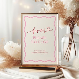 May include: A gold-framed sign with the word "favors" in pink script, a pink heart, and the text "PLEASE TAKE ONE." The sign is on a gold stand, with a marble surface and floral arrangement in the background.