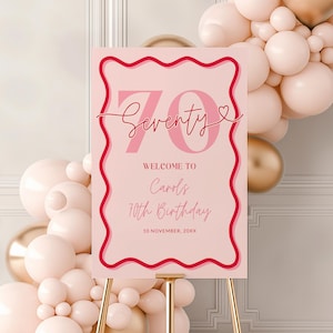 May include: A pink and red birthday sign on a gold easel. The sign reads "Seventy" with the text "Welcome to Carols 70th Birthday 10 November, 20XX". The background features a balloon arch in shades of pink and gold.