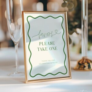 May include: A gold framed sign with a white background and green wavy border. The sign reads "favors, please take one" in elegant script. The bottom of the sign says "Thank you for celebrating with us!"
