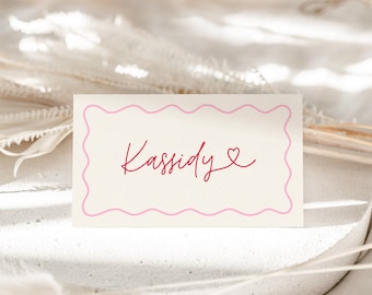 May include: A white place card with a pink wavy border. The name "Kassidy" is written in red cursive with a small red heart next to it.