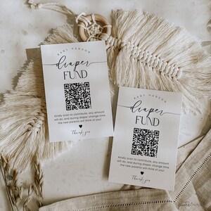 Könnte beinhalten: Zwei weiße Karten mit schwarzem Text und einem schwarz-weißen QR-Code. Der Text auf den Karten lautet "Baby Hanson Diaper Fund" und enthält Anweisungen, den QR-Code zu scannen, um zum Windelfonds beizutragen. Die Karten liegen auf einer hellbraunen Oberfläche mit einem beigefarbenen Makramee-Hänger im Hintergrund.