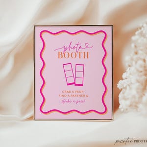 May include: A framed pink photo booth sign with a wavy magenta border and gold accents. The sign reads "photo booth" in cursive, with the words "Grab a prop, find a partner & strike a pose!" below.