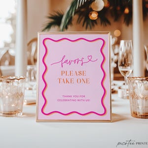May include: A framed pink sign with gold trim and text that reads "favors, PLEASE TAKE ONE, THANK YOU FOR CELEBRATING WITH US!" The sign is on a white table with candles and glasses in the background.