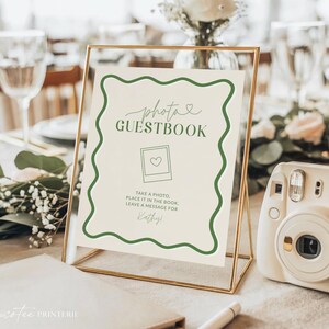 Könnte beinhalten: Goldgerahmtes Schild mit den Worten "photo GUESTBOOK" in grüner Schrift. Das Schild hat einen grünen Wellenrand und Anweisungen. Rechts befindet sich eine weiße Sofortbildkamera.