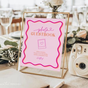 Könnte beinhalten: Ein rosa Foto-Gästebuch-Schild in einem goldenen Rahmen mit dem Text "photo GUESTBOOK". Das Schild enthält eine Polaroid-Grafik mit einem Herzen und Anweisungen. Eine weiße Sofortbildkamera befindet sich daneben.