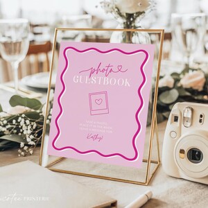 Może przedstawiać: Różowy znak księgi gości ze zdjęciami w złotej ramie z napisem "photo GUESTBOOK". Znak ma falistą różową obwódkę i grafikę polaroid z sercem. Po prawej stronie znajduje się biały aparat natychmiastowy.