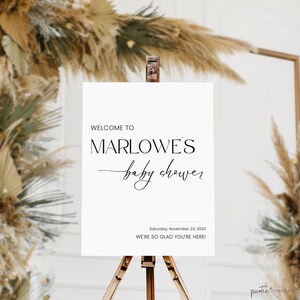 May include: A white sign on a wooden easel with the text "WELCOME TO MARLOWE'S baby shower" in black script. The date "Saturday, November 23, 2023" and the phrase "WE'RE SO GLAD YOU'RE HERE!" are also displayed.