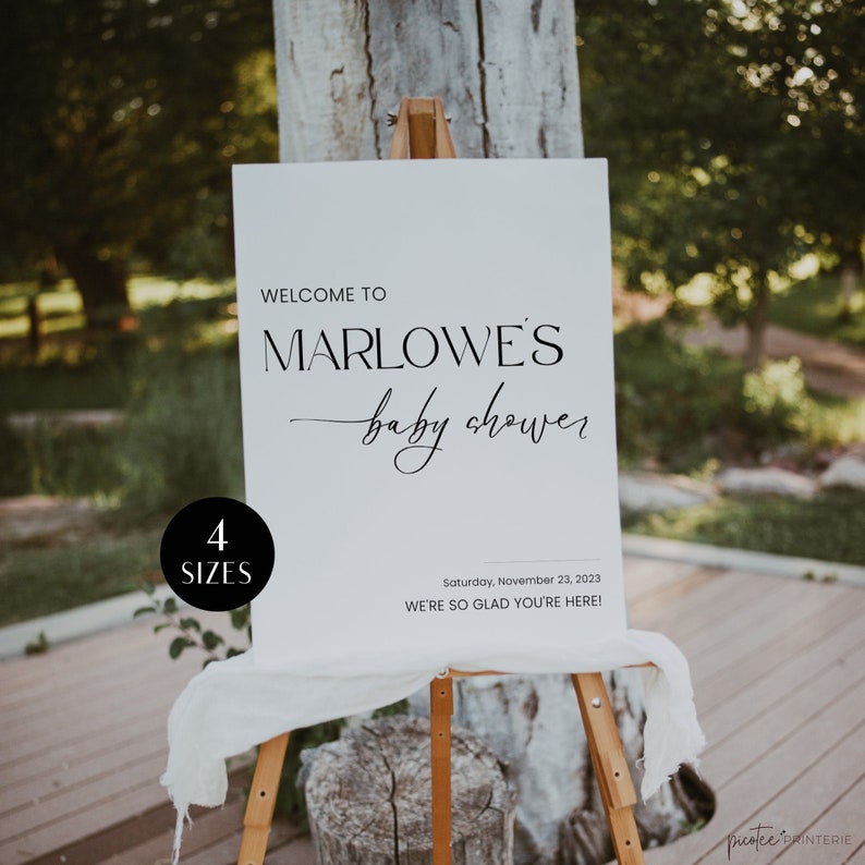 May include: A white sign on a wooden easel with the text "WELCOME TO MARLOWE'S baby shower." The sign also includes the date "Saturday, November 23, 2023" and the phrase "WE'RE SO GLAD YOU'RE HERE!" A black circle with the text "4 SIZES" is in the lower left corner.