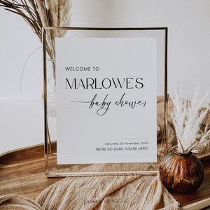 May include: A framed white sign with gold edging, announcing a baby shower. The text reads "WELCOME TO MARLOWE'S baby shower" in black. The date and a welcoming message are also included. The sign is surrounded by decorative elements.