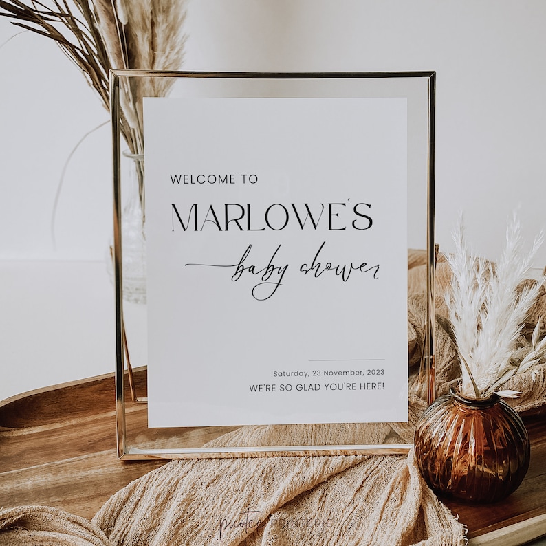May include: A framed white sign with gold edging, announcing a baby shower. The text reads "WELCOME TO MARLOWE'S baby shower" in black. The date and a welcoming message are also included. The sign is surrounded by decorative elements.