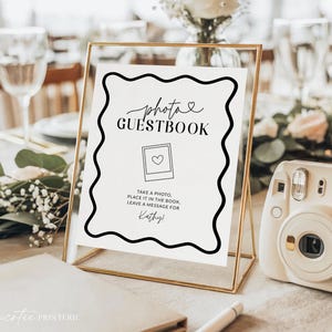 Może przedstawiać: Złota ramka z napisem "photo GUESTBOOK" elegancką czcionką, z sercem i ikoną polaroidu. Znak zawiera instrukcje, aby zrobić zdjęcie, umieścić je w książce i zostawić wiadomość dla Kathy. W pobliżu znajduje się biały aparat natychmiastowy.