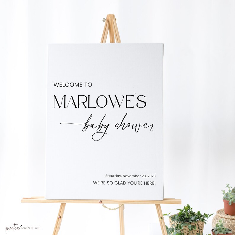 May include: A white sign on a wooden easel with the text "WELCOME TO MARLOWE'S baby shower" in black. Below, it reads "Saturday, November 23, 2023 WE'RE SO GLAD YOU'RE HERE!" The sign is for a baby shower.