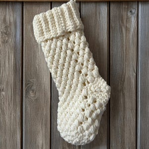 May include: A cream-coloured, handmade Christmas stocking. The stocking is made of a thick, textured yarn with a bobble stitch pattern. The top of the stocking is folded over, and a loop is attached for hanging. The background is a wooden wall.