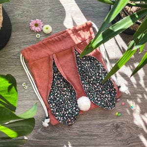 May include: A coral corduroy drawstring bag with floral bunny ears and white pom-pom tails. The bag is on a wooden surface, with small flowers and greenery in the background.