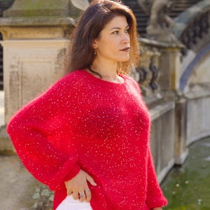 May include: A woman wearing a red sweater with sequins and white pants. She is standing in front of a stone fountain.